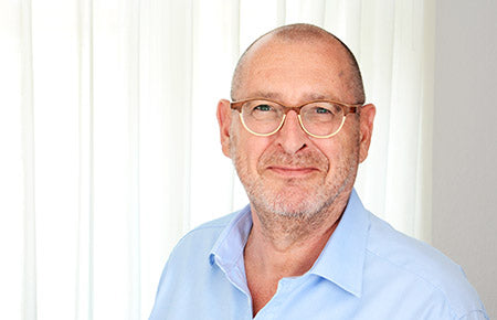 Ein Mann mittleren Alters mit Brille und hellblauem Hemd lächelt warm vor weißen Vorhängen, bereit, Erkenntnisse aus "Hinter verschlossenen Türen: Psychiater Erzählungen von Drs. Dolf Hage" von ShapeMinds. Schulungen und Handouts für Mitarbeiter