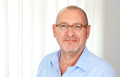 Ein Mann mittleren Alters mit Brille und hellblauem Hemd lächelt warm vor weißen Vorhängen, bereit, Erkenntnisse aus "Hinter verschlossenen Türen: Psychiater Erzählungen von Drs. Dolf Hage" von ShapeMinds. Schulungen und Handouts für Mitarbeiter